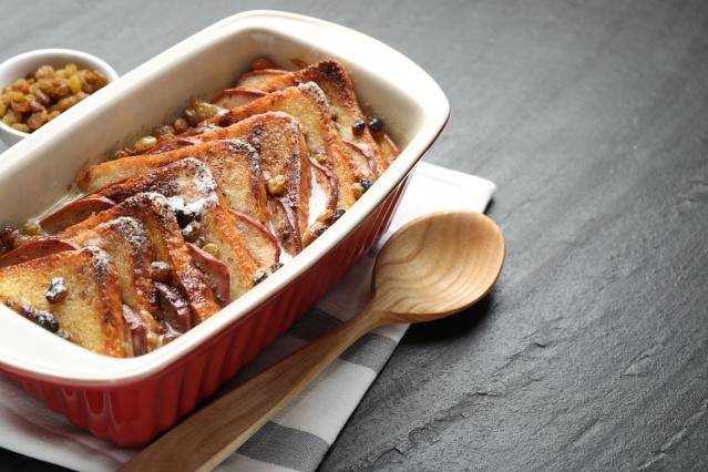 A baked bread pudding with sliced fruit and raisins sits in a red baking dish dusted with icing sugar. A wooden spoon rests beside the dish on a dark tabletop.