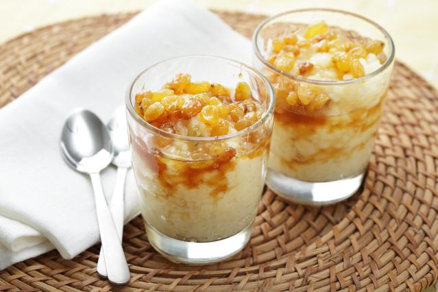 Two glasses of rice pudding topped with raisins and fruit sauce sit on a woven placemat beside 2 teaspoons and a folded white napkin.