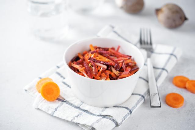 A small bowl of grated beetroot, carrot, and apple salad on a white cloth beside sliced carrots and cutlery.