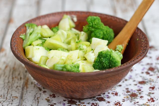 A wooden bowl filled with mixed green vegetables, including broccoli and chopped pale green vegetables, with a wooden spoon resting inside the bowl.