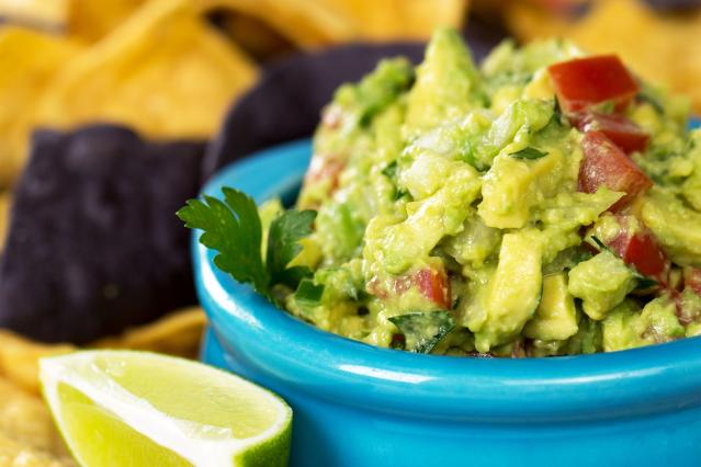 A blue bowl filled with chunky guacamole made with avocado and tomato, served with corn chips and a lime wedge nearby.