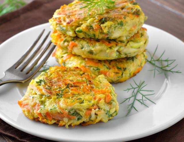 A stack of golden vegetable fritters with visible grated carrot and herbs sits on a white plate beside a fork.