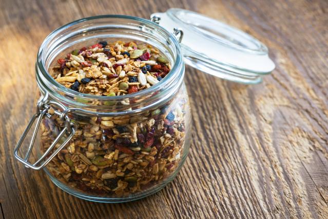 A glass jar filled with homemade toasted muesli containing oats, seeds, nuts and dried fruit sits open on a wooden table.