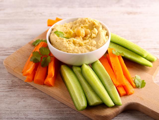 A bowl of hummus served with carrot and cucumber sticks on a wooden board.