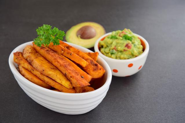 A bowl of roasted kūmara chips seasoned with spices, served with a small bowl of chunky guacamole. Half an avocado and parsley garnish sit in the background.