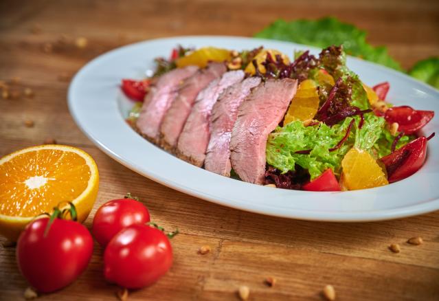 Sliced lamb served on a salad with leafy greens, orange segments, cherry tomatoes and kūmara on a white plate.