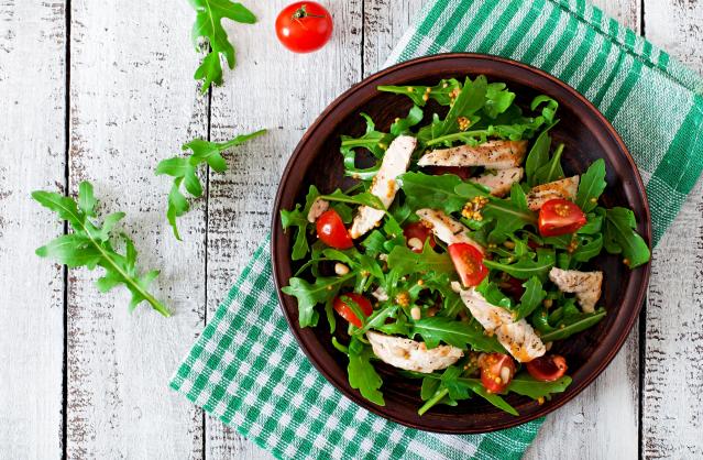 A bowl of chicken and rocket salad with cherry tomatoes and dressing on a wooden table beside a green checked cloth.