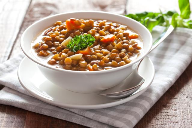 A bowl of lentil and vegetable soup with carrots and herbs, served on a white plate with a spoon on a wooden table.