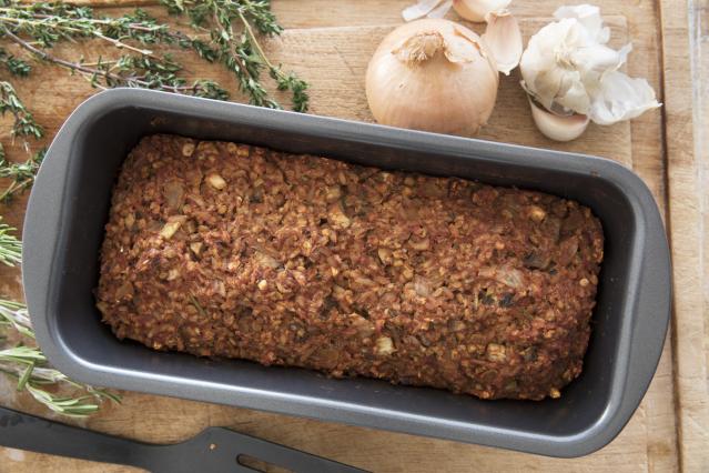 A homemade meat loaf in a loaf tin, surrounded by garlic, onion and fresh herbs on a wooden board.
