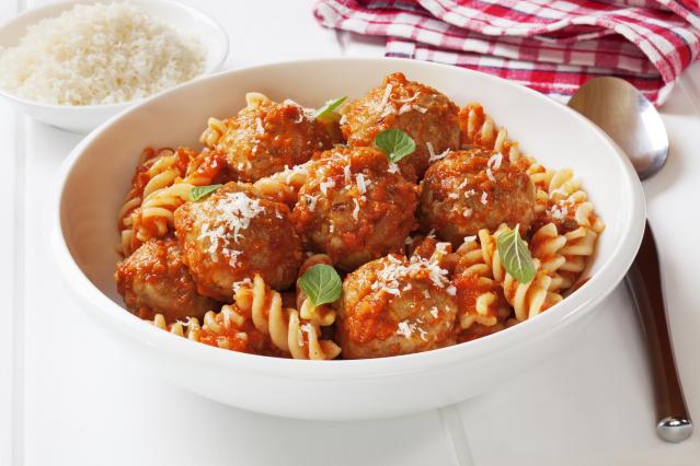 A bowl of meatballs in tomato sauce served with spiral pasta and grated cheese, with a bowl of grated parmesan in the background.