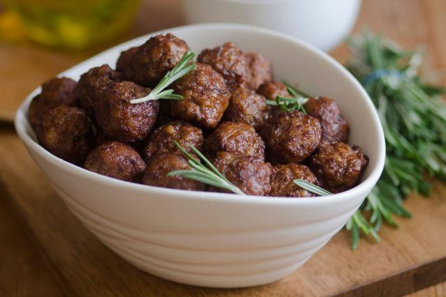 A bowl of cooked meatballs garnished with rosemary on a wooden board.