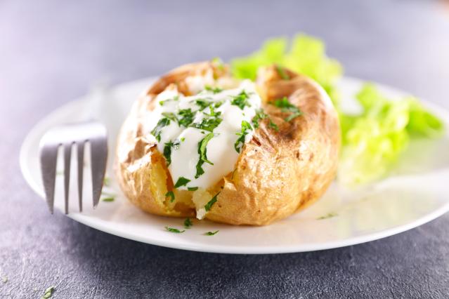 A baked potato topped with sour cream and chopped herbs on a white plate.