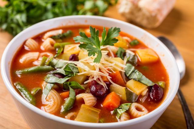 A bowl of minestrone soup with pasta, beans, green beans, carrot, courgette, and herbs in a tomato-based broth, topped with grated cheese and parsley.