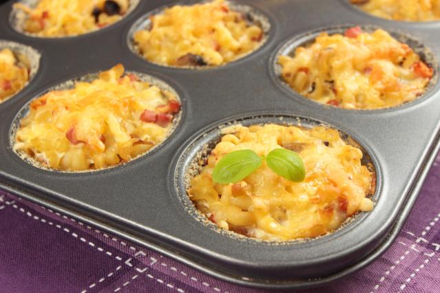 Mini cheesy pasta bakes cooked in a muffin tray, topped with melted cheese and basil leaves.