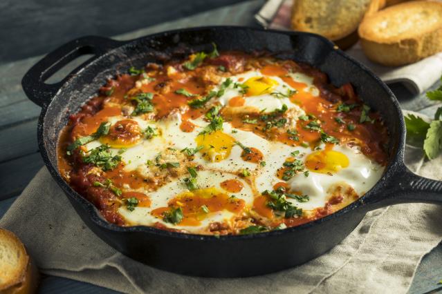 A cast-iron pan filled with baked eggs in a rich tomato sauce, topped with herbs, served with slices of bread on the side.