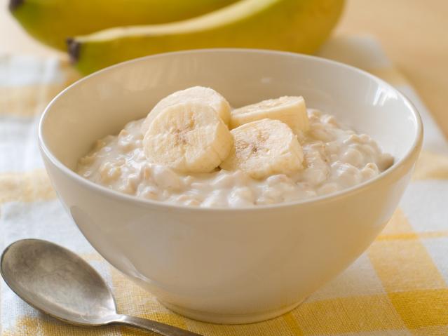 A bowl of creamy porridge topped with sliced banana, with a spoon beside the bowl and bananas in the background.