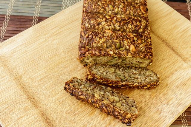 A loaf of seeded oat bread on a wooden board, with slices cut to show the dense texture filled with oats, seeds and nuts.