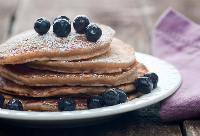 A stack of oat pancakes topped with fresh blueberries and a dusting of icing sugar on a white plate.