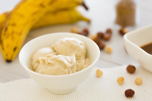 A bowl of creamy banana ice cream with bananas and scattered nuts in the background.