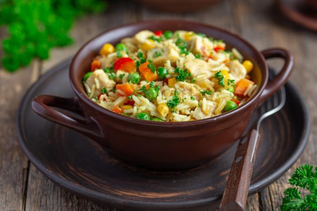 A bowl of one-pot chicken and rice with mixed vegetables including peas, corn, carrots and capsicum, topped with chopped parsley.