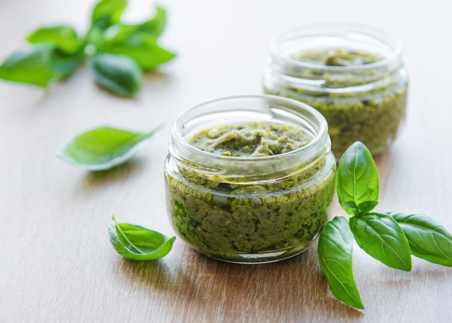Two small glass jars of green pesto with fresh basil leaves on a wooden table.