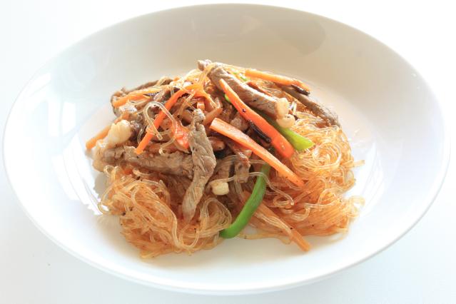 A bowl of stir-fried glass noodles with sliced pork and vegetables including carrot and green capsicum.