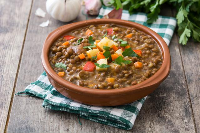 A bowl of pork, lentil and vegetable stew topped with chopped parsley on a wooden table.