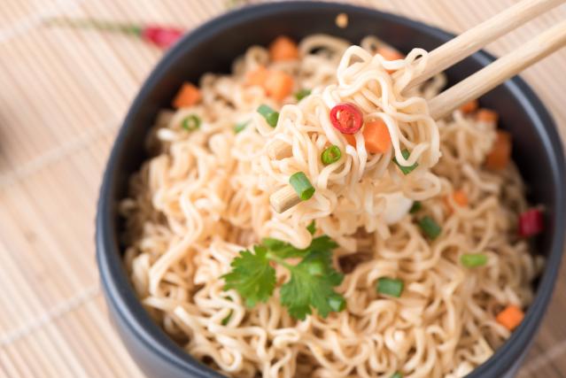 A bowl of quick noodles with chopped vegetables and herbs, being lifted with chopsticks.