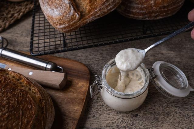 Jar of bubbly rewena bread starter being scooped with a spoon beside freshly baked loaves of bread.