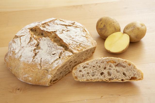 Loaf of rewena bread with a sliced piece beside it, next to whole and halved potatoes on a wooden table.