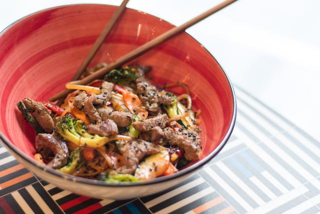 Bowl of sapasui-style stir-fried noodles with beef and vegetables, topped with black sesame seeds and served with chopsticks.