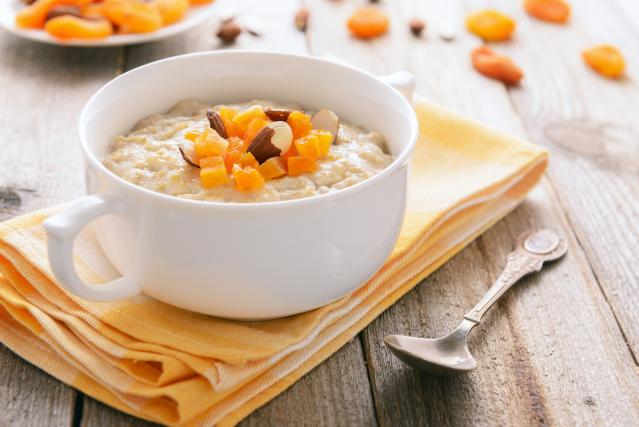 A bowl of creamy porridge topped with chopped dried apricots and almonds, sitting on a yellow cloth beside a spoon.