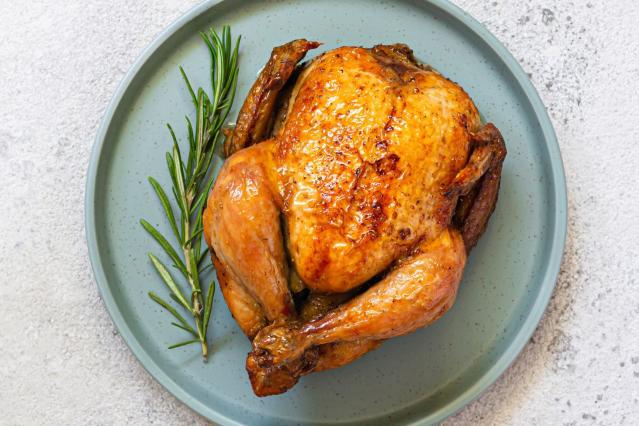 A whole roast chicken with crispy golden skin on a plate beside a sprig of rosemary.