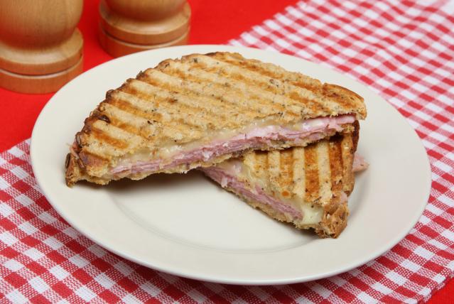 Two halves of a toasted ham and melted cheese sandwich with grill marks on a white plate on a red checked tablecloth.