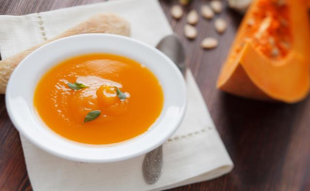 A bowl of bright orange pumpkin soup garnished with cream and small green leaves, sitting on a cloth napkin beside a spoon and bread roll.