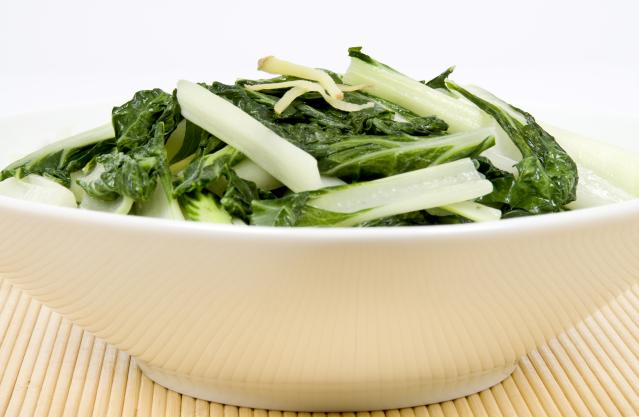 A bowl of cooked bok choy with leafy greens and pale stems, topped with thin slices of ginger.