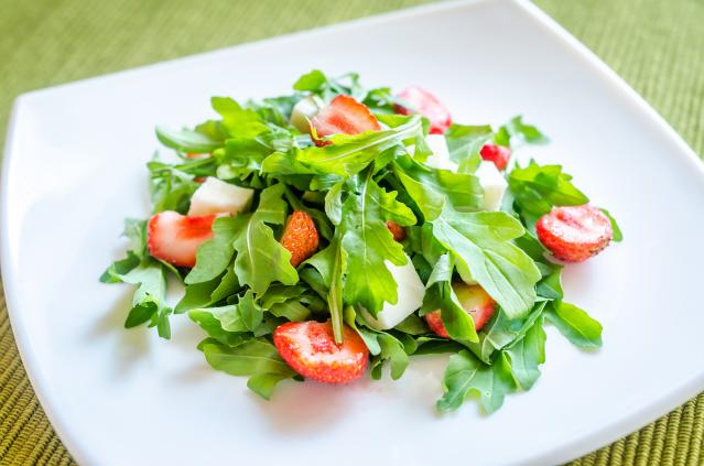 A fresh salad made with leafy greens, sliced strawberries, and cubes of white cheese on a square white plate.