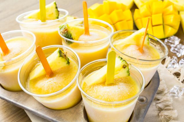 Homemade tropical ice blocks in small cups with wooden sticks and pieces of pineapple on top.