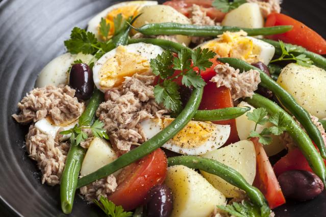 Tuna salad with green beans, boiled eggs, potatoes, tomatoes, olives, and parsley in a dark bowl.