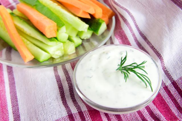 Bowl of tzatziki dip beside carrot and cucumber sticks on a striped cloth.