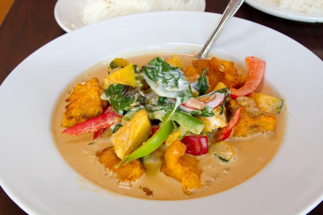 Vegetable curry with tofu, capsicum, leafy greens, and vegetables in a creamy sauce served in a white bowl with rice in the background.