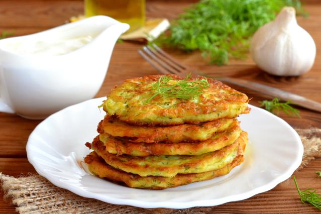 Stack of golden vegetable pikelets garnished with dill on a white plate, with garlic and sauce in the background.