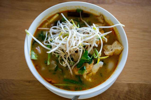 Bowl of vegetable tom yum soup topped with bean sprouts, herbs and vegetables on a wooden table.