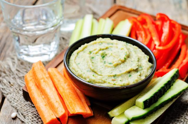 Bowl of creamy green hummus served with sliced carrot, cucumber, celery and red capsicum sticks on a wooden board.
