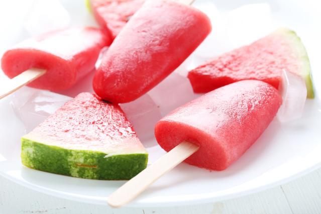 Homemade watermelon ice blocks on sticks served on a white plate with fresh watermelon slices and ice cubes.
