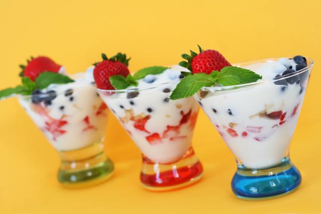 Three glass cups filled with yoghurt, strawberries, blueberries, and cereal or granola, topped with fresh strawberries and mint leaves, against a bright yellow background.