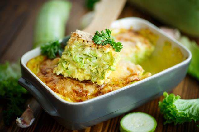 A baked zucchini slice in a grey baking dish with a piece lifted out on a wooden spatula. The slice has layers of zucchini, egg, and melted cheese, and is topped with parsley. Fresh zucchini and lettuce are blurred in the background.