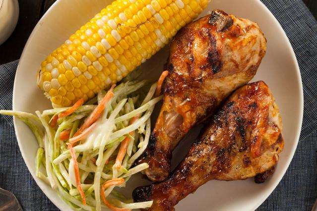 A plate containing two grilled chicken drumsticks, a cob of corn, and creamy coleslaw made with shredded cabbage and carrot. The chicken is golden brown with charred edges.
