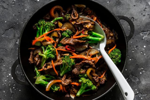 A pan of beef and broccoli stir-fry with sliced beef, broccoli florets, carrot strips, onions, and capsicum in a dark sauce. A serving spoon rests in the pan against a dark background.
