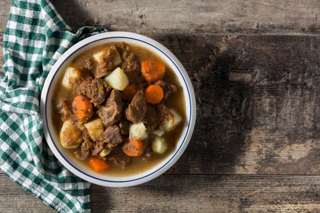 A bowl of beef and vegetable stew containing chunks of beef, potato, and carrot in a rich gravy. The bowl sits on a rustic wooden table beside a green and white checked tea towel.